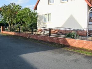 Metallzaun an Wohnhaus mit Ziegelsockel und Grünanlage.