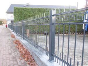 Metallzaun an Straße mit Hecke und Herbstlaub.