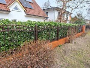 Brauner Schmiedezaun mit dekorativen Elementen, montiert auf roter Ziegelmauer vor einem Einfamilienhaus