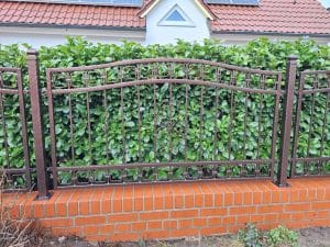 Brauner Schmiedeeisenzaun mit dekorativen Elementen, montiert auf einer niedrigen roten Klinkermauer vor dichtem Grün