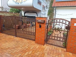 Braunes Schmiedeeisentor mit Tür und Initialen AJK, montiert zwischen roten Ziegelpfeilern vor einem Haus mit Wohnmobil