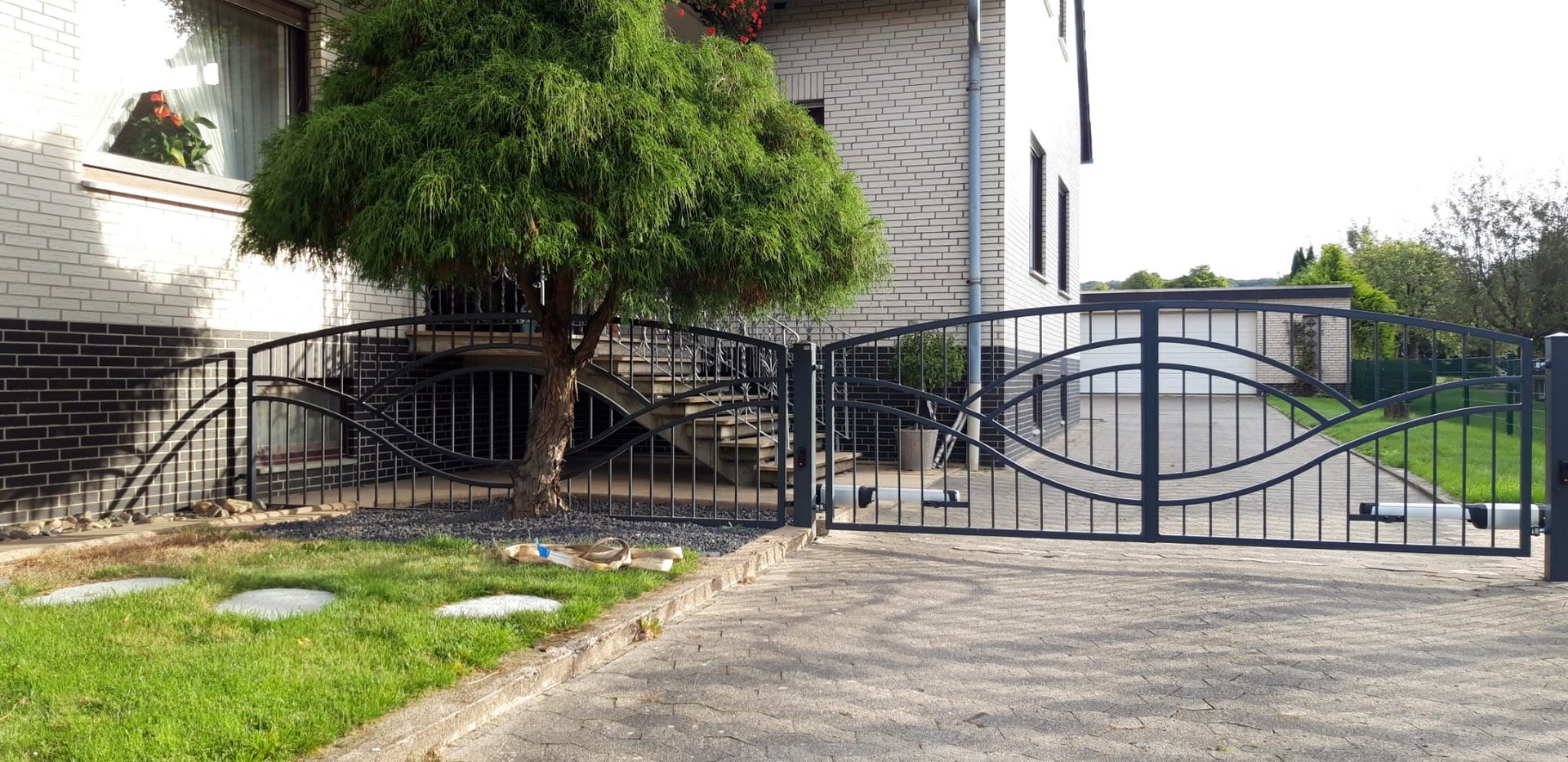 Einfahrt mit geschwungenem Metalltor und Bäumen.